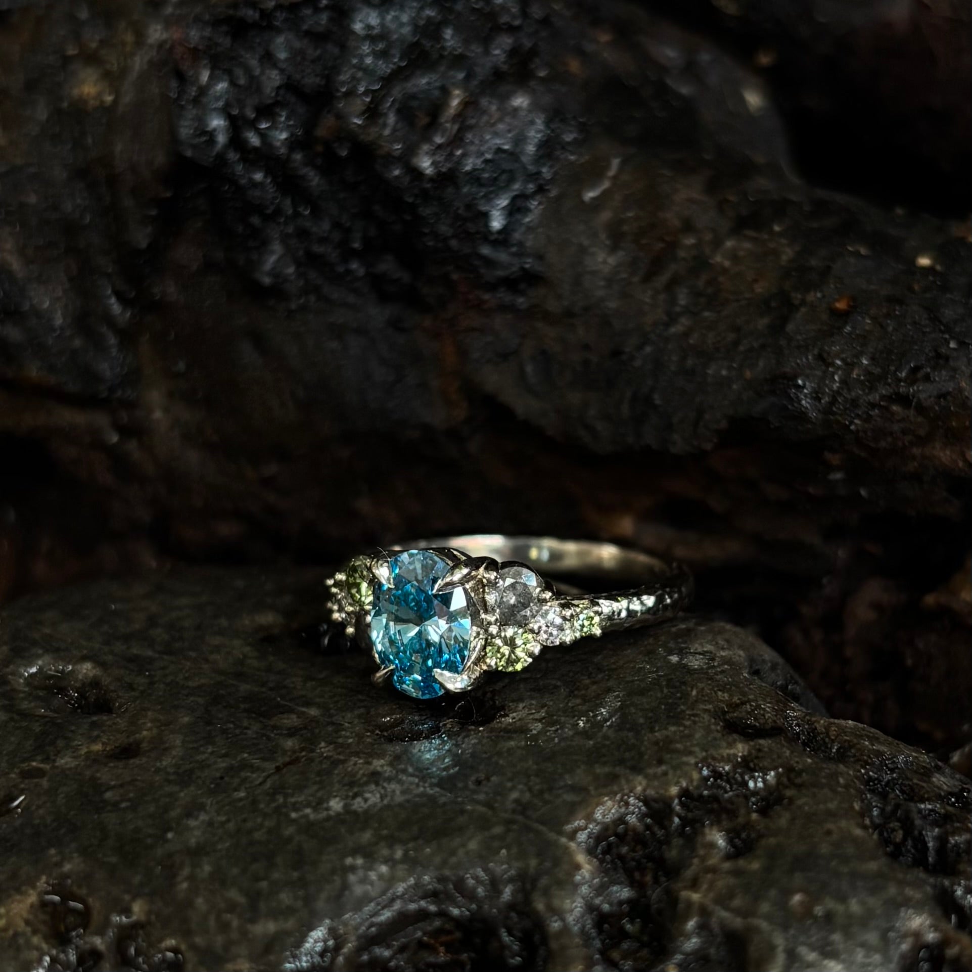 3/4 view White gold Ring with a central oval blue gemstone and grey, green and white accent stones on a dark stone surface