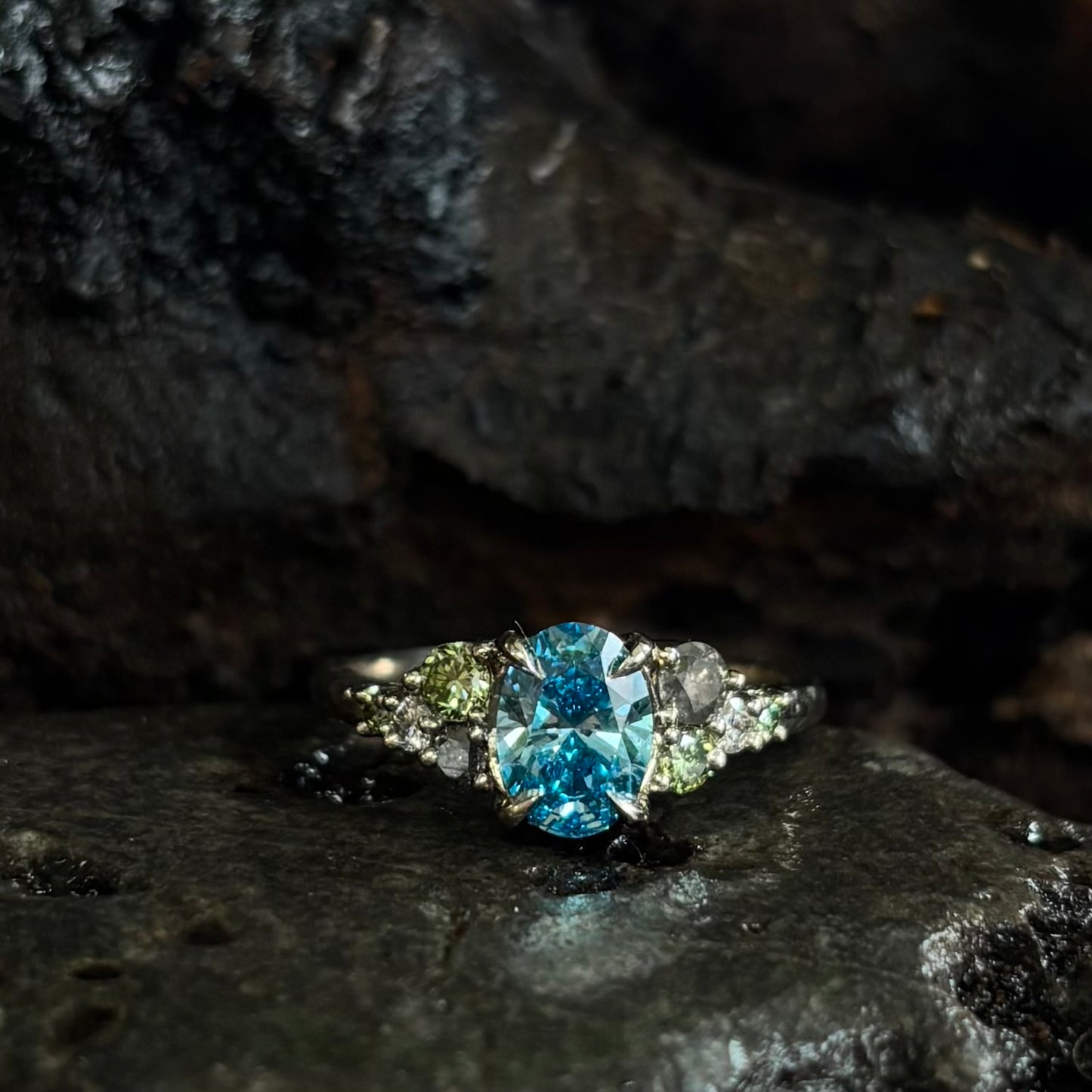 White gold Ring with a central oval blue gemstone and grey, green and white accent stones on a dark stone surface