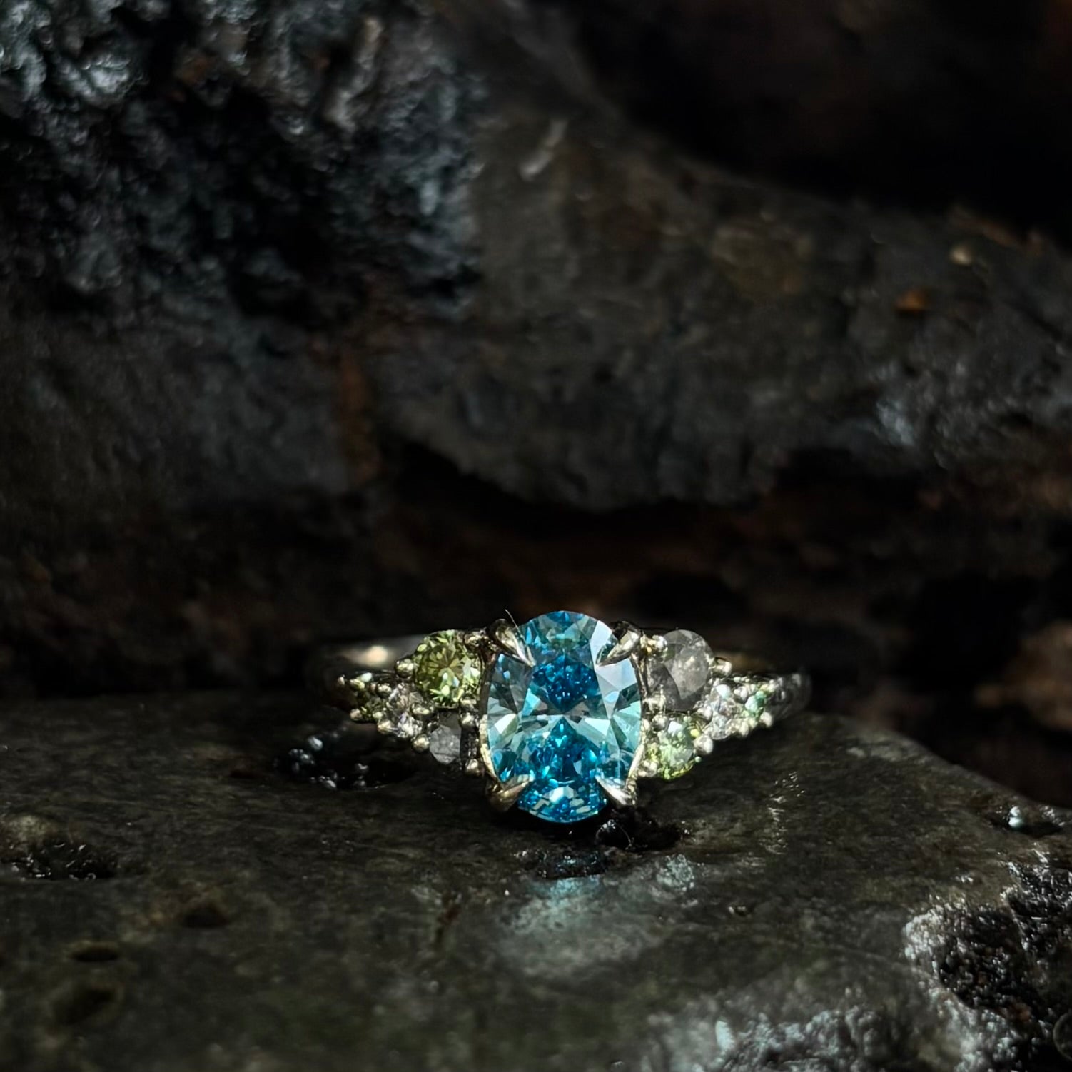 White gold Ring with a central oval blue gemstone and grey, green and white accent stones on a dark stone surface
