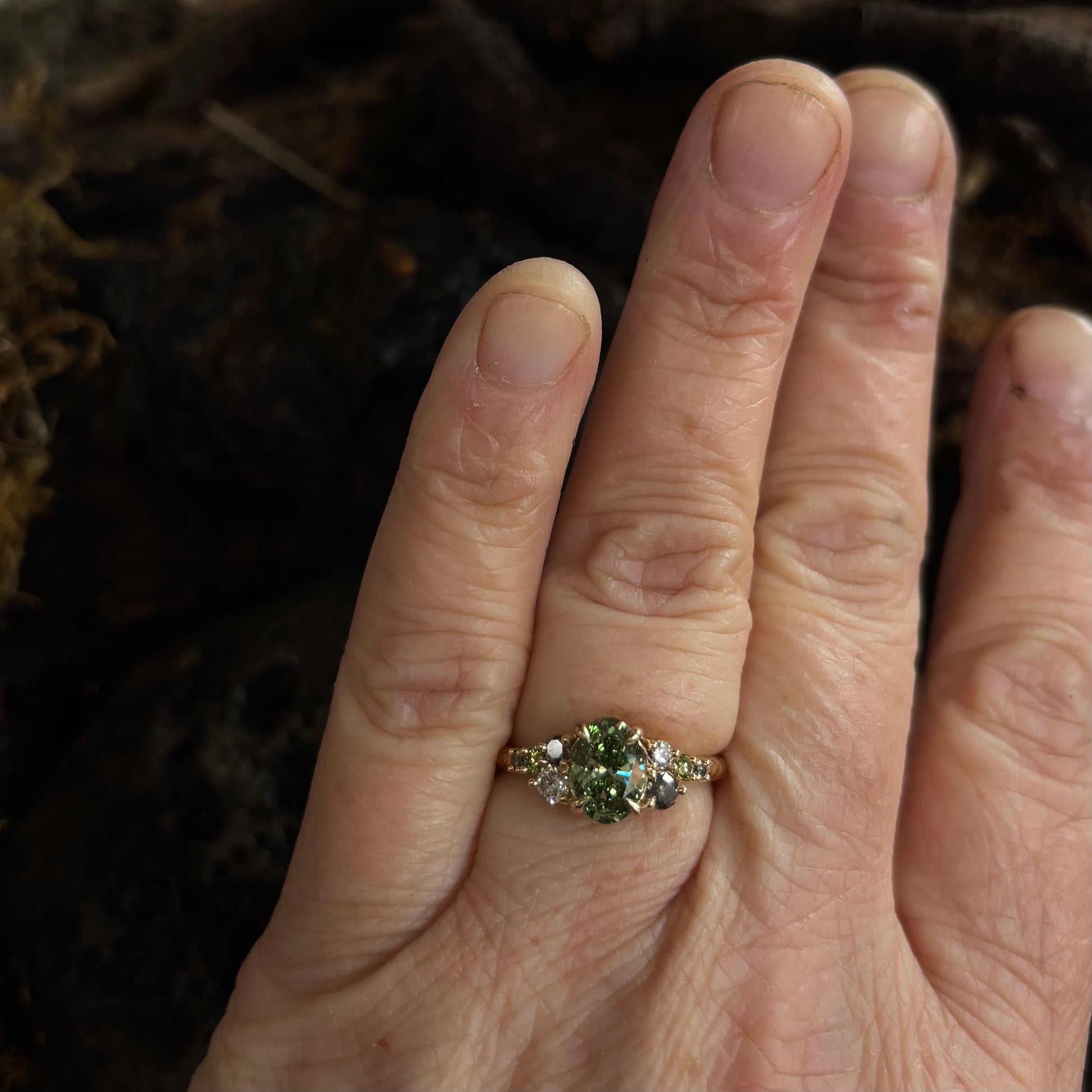 Hand wearing a Ring with a Central green gemstone and grey green and white accent stones on a dark textured surface