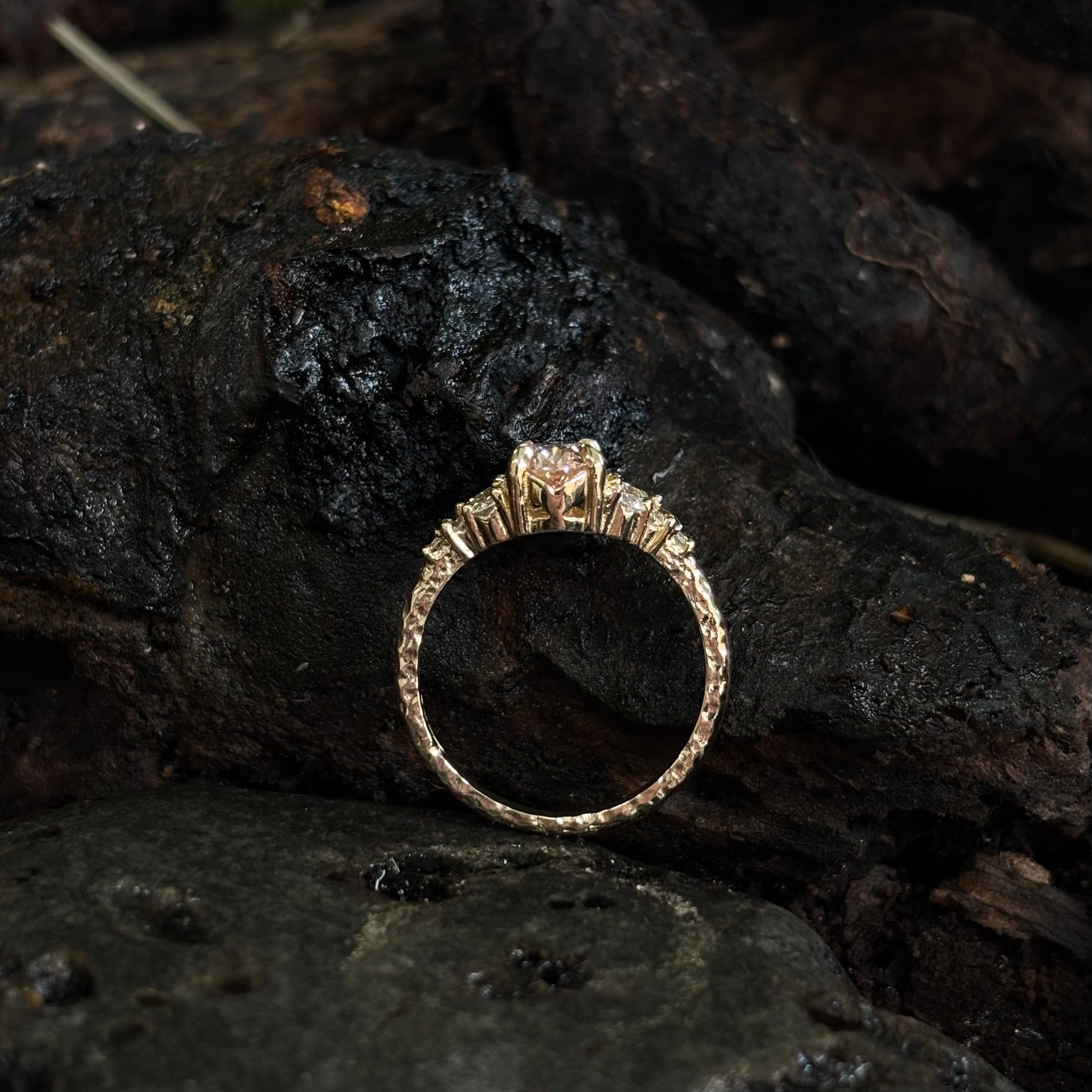 Oval pink diamond with white diamond side stones in yellow gold on dark Textured surface