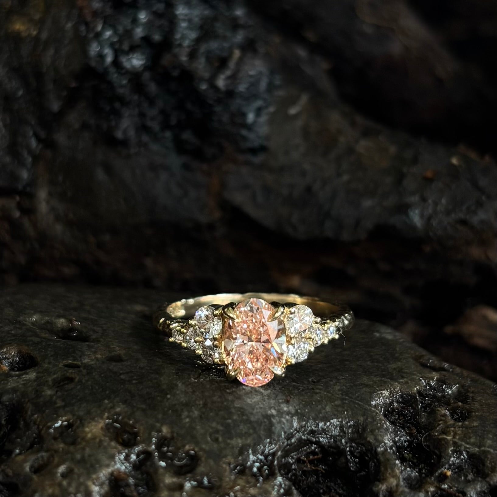 Front view of Oval pink diamond with white diamond side stones in yellow gold on dark background